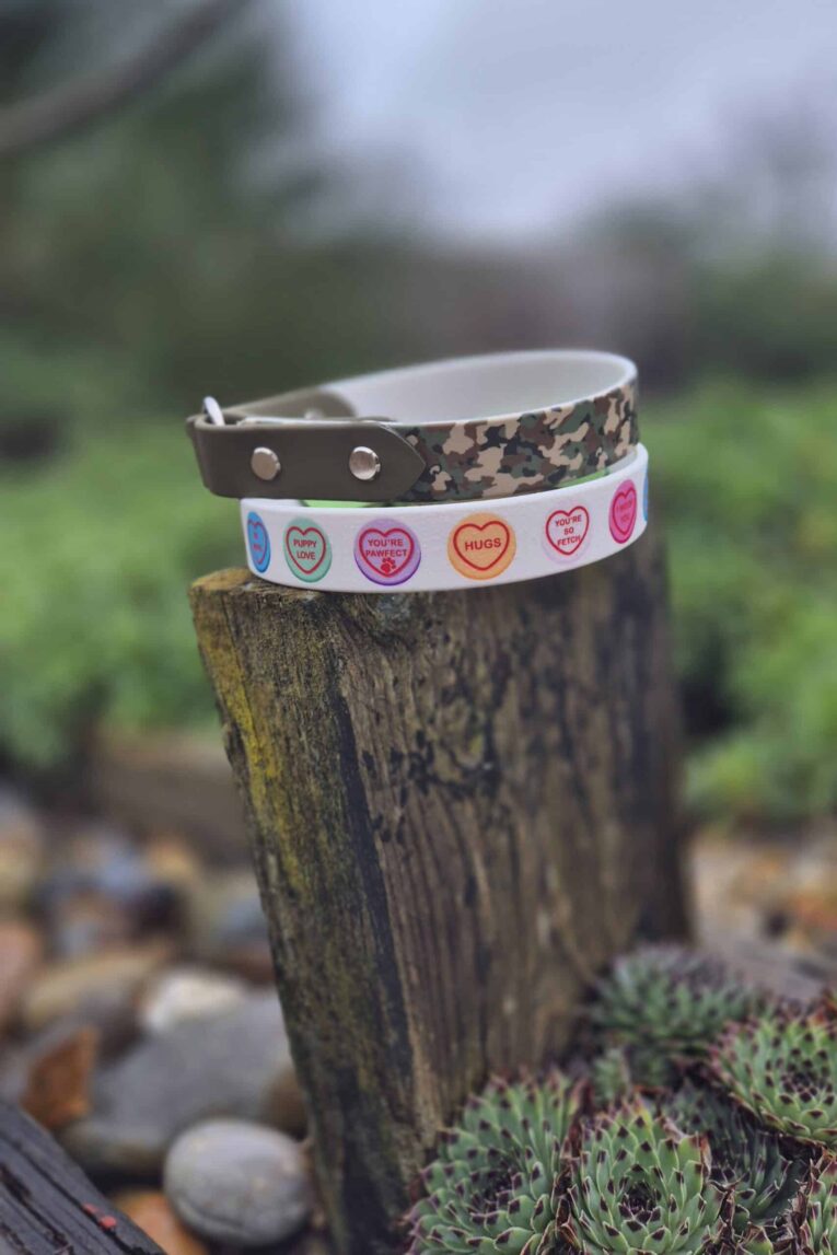Two handmade BioThane dog collars stacked on weathered wood: camouflage design with studs and white printed collar with colourful heart motifs.
