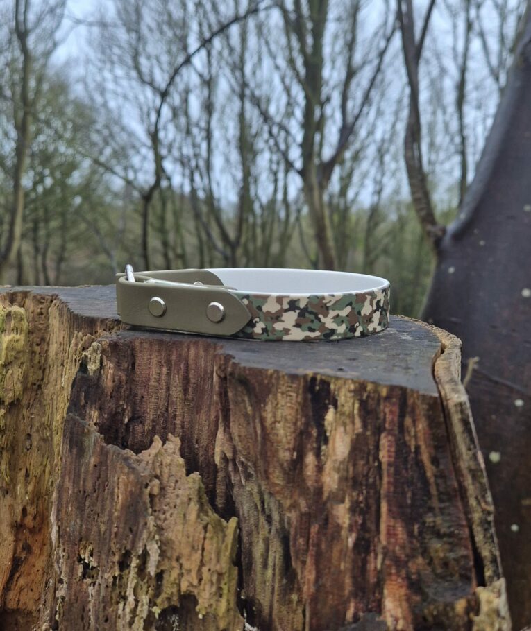 Handmade BioThane dog tag collar in camouflage print with silver hardware, displayed on weathered wood.