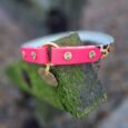 Vibrant pink handmade BioThane tag collar with gold studs and ring, displayed on moss-covered wood.