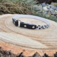 Handmade BioThane tag collar with black and white cow-print design and metal fixings, displayed on wood.