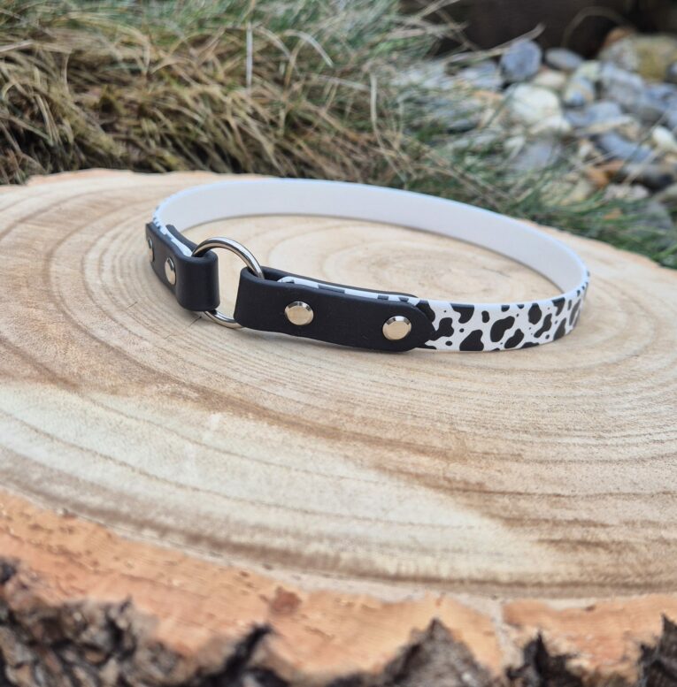 Handmade BioThane tag collar with black and white cow-print design and metal fixings, displayed on wood.