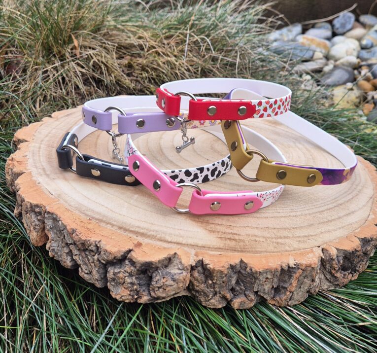 Colourful handmade BioThane tag collars displayed on wood, featuring red hearts, pink, purple, black and patterned designs with metal hardware.