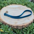 Bright blue BioThane traffic handle lead with brass fittings, displayed on wooden disc surrounded by green foliage.