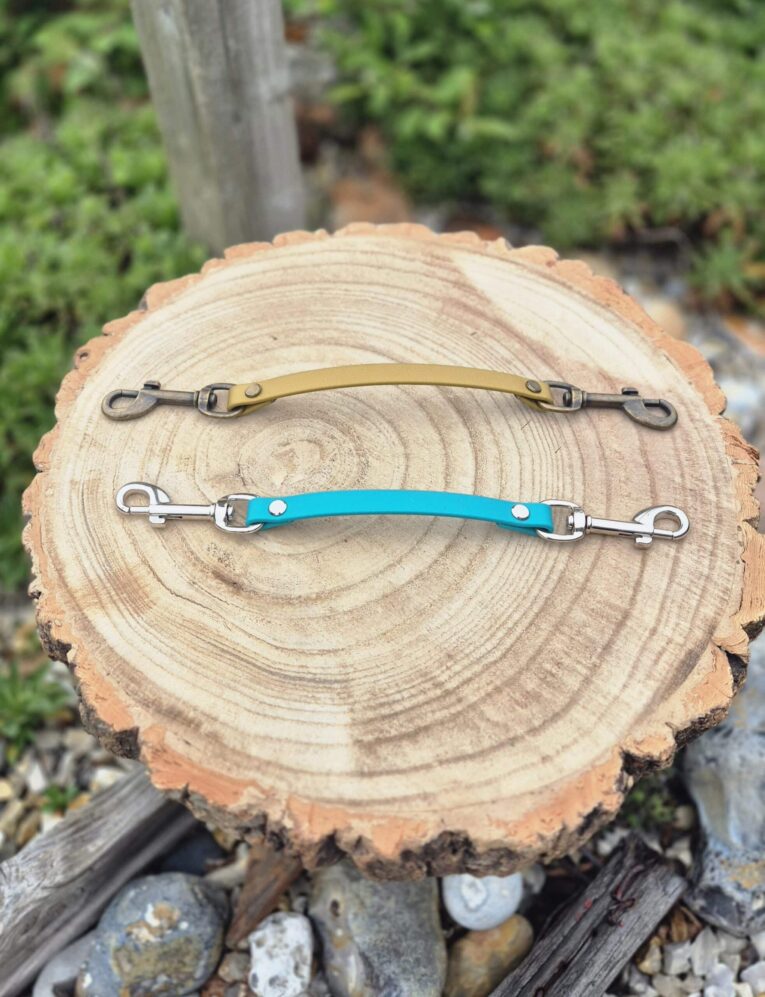 Two handmade BioThane dog collar safety clips with metal hardware displayed on a wooden log cross-section.