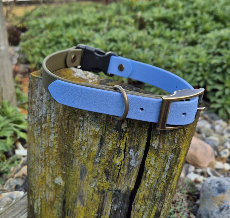 Two handmade BioThane dog collars in blue and sage green, displayed on a weathered wooden post outdoors.