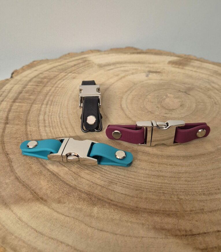 Three handmade BioThane quick release buckles in black, burgundy, and turquoise displayed on a wooden cross-section.