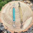 Two BioThane safety clips in turquoise and tan displayed on a wooden log cross-section.