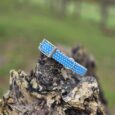 Blue polka dot handmade BioThane dog collar displayed on textured bark with rural landscape background.