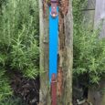 Blue tapered handmade BioThane dog collar with brown hardware, displayed on weathered wood post surrounded by greenery.