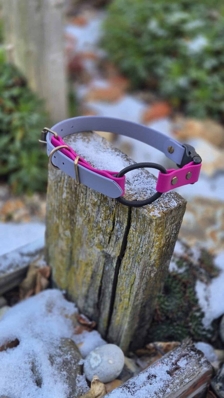 Handmade BioThane dog collar in pink and grey with black ring, displayed on weathered wood.