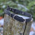 Two black BioThane dog collars with metal D-rings and buckles displayed on weathered wood outdoors.
