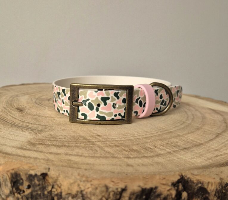 Handmade BioThane dog collar with camouflage print pattern, brass buckle and pink detailing on wooden surface.