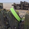 Bright lime-green double-layered dog collar displayed on weathered wooden post by beach with historic pier ruins blurred behind.