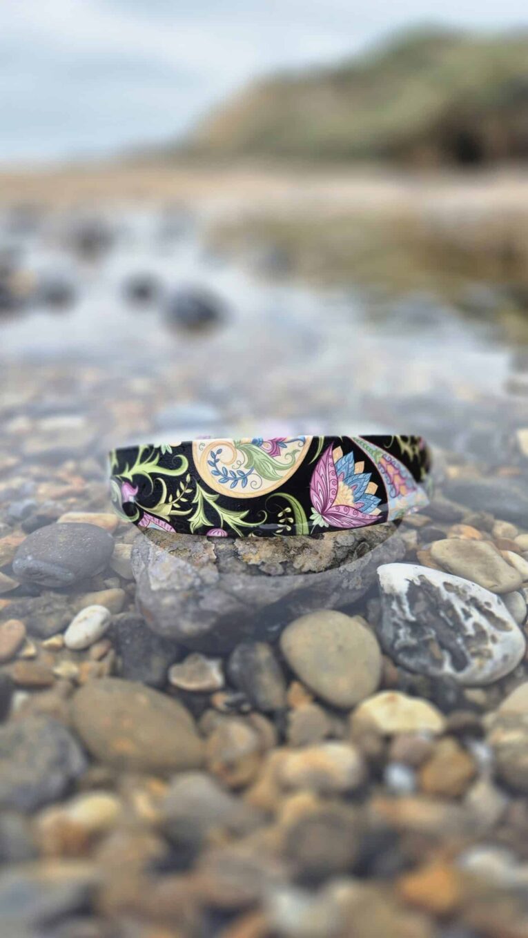 Handmade BioThane dog collar with floral waterproof design, positioned on pebbles by the shore.