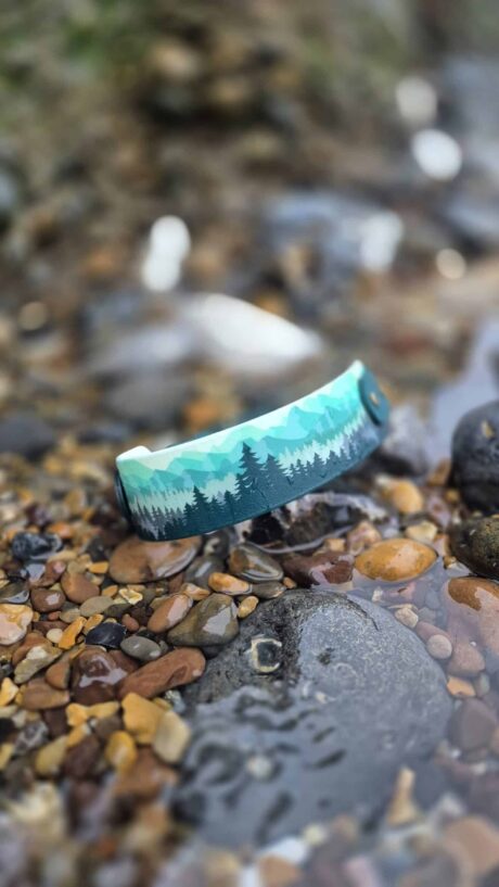 Handmade forest print dog collar positioned on pebbles and stones, showcasing turquoise woodland design with layered evergreen trees.