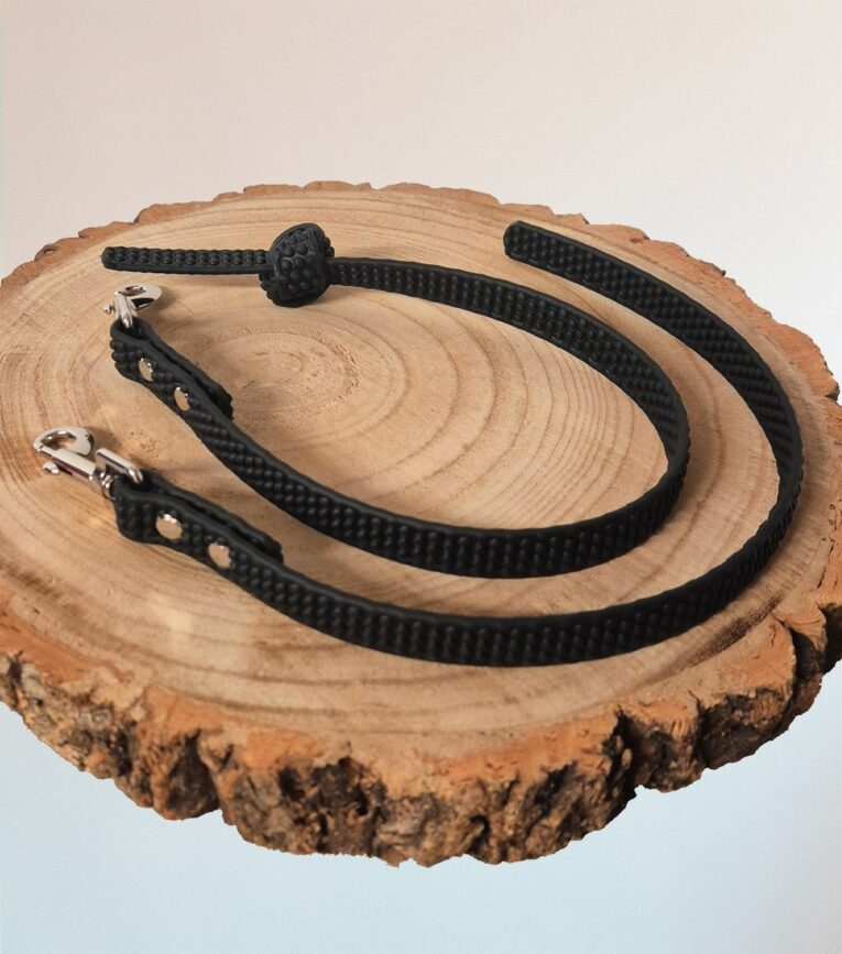 Black handmade BioThane dog collars and leads displayed on natural wood, featuring metal hardware and grip sections.