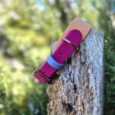 Brightly coloured handmade BioThane dog collar in magenta and blue, displayed on weathered wood with garden foliage behind.