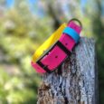 Colourful handmade BioThane dog collars stacked on weathered wood with blurred foliage background.