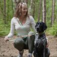 Woman crouching with black and white dog wearing printed BioThane collar and lead in woodland setting.
