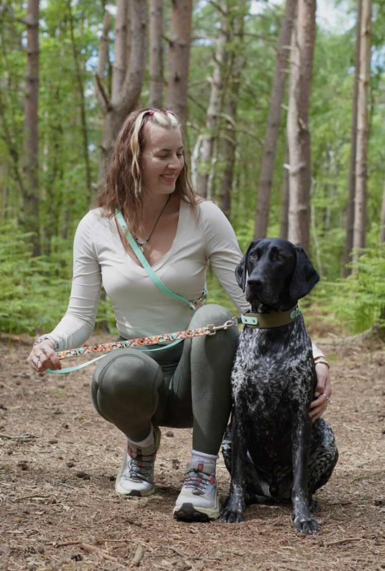 Woman crouching with black and white dog wearing printed BioThane collar and lead in woodland setting.