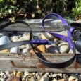 Midnight amethyst handmade BioThane dog collars and leads displayed on weathered wood with stones and foliage.