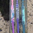 Three handmade BioThane dog collars with patterned designs in pink, purple, and teal, displayed on weathered wood.