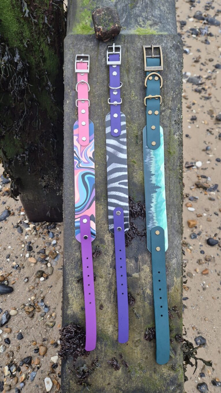 Three handmade BioThane dog collars with patterned designs in pink, purple, and teal, displayed on weathered wood.