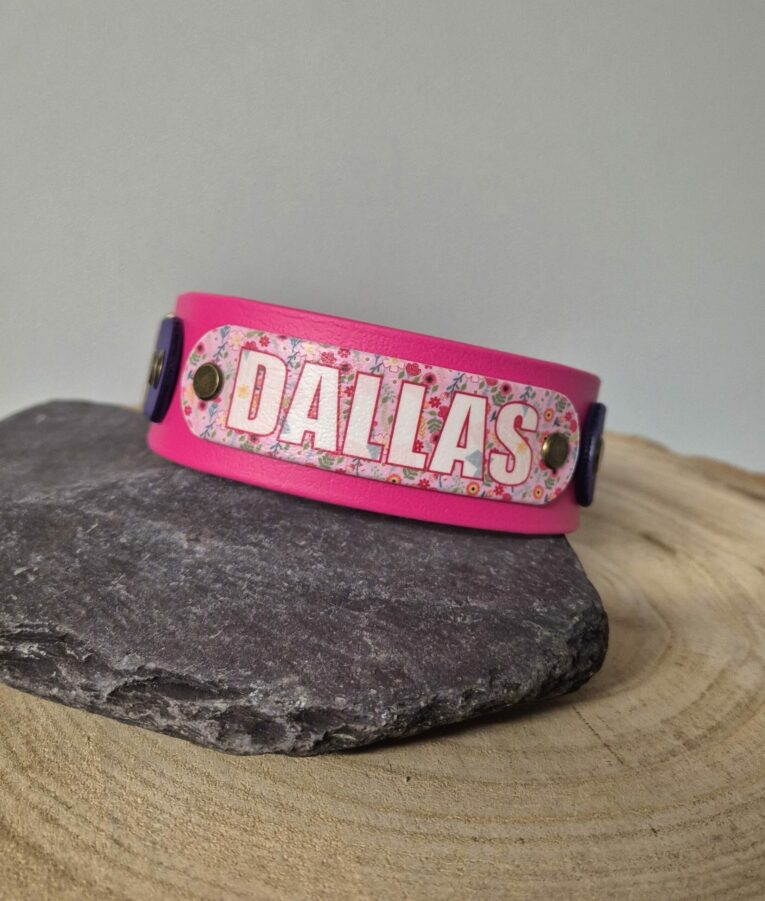 Bright pink personalised dog collar with floral printed panel reading 'DALLAS' on grey stone and wooden surface.