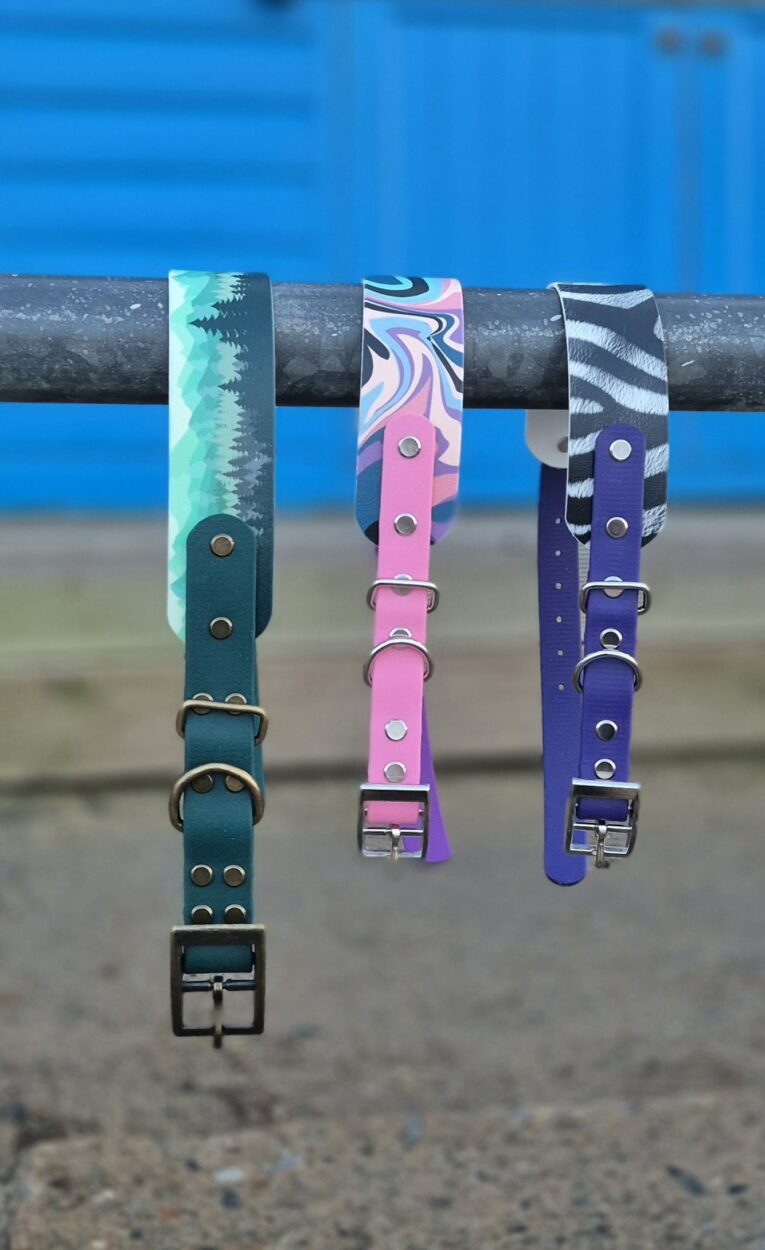 Three handmade BioThane dog collars with printed designs displayed on a seaside rail; turquoise, pink, and purple styles.