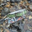 Two handmade BioThane dog collars displayed on coastal rocks: one with lime green diamond print, one with koi fish artwork in orange and blue.
