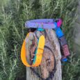 Colourful handmade BioThane dog collars and leads displayed on weathered wood post with greenery behind.
