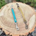 Two safety clips for dog collars in turquoise and olive, displayed on a wood cross-section.