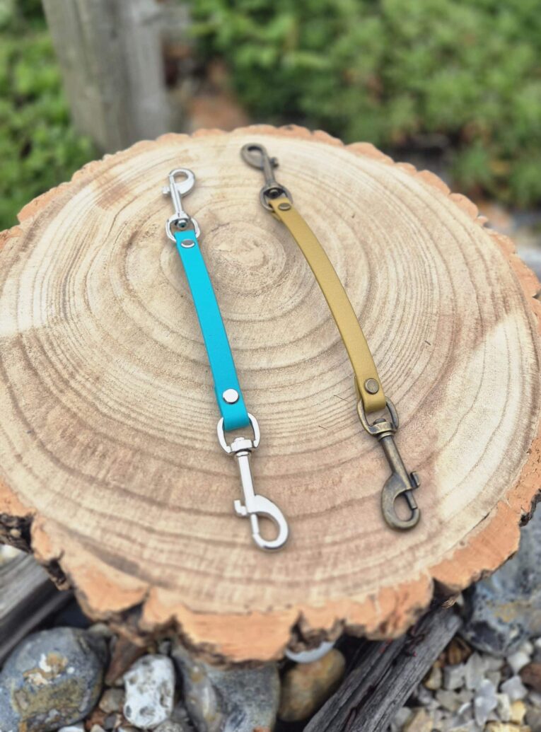 Two safety clips for dog collars in turquoise and olive, displayed on a wood cross-section.