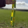 Waterproof gundog collar in yellow and green BioThane, held aloft against a countryside sky.
