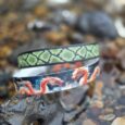 Three handmade BioThane dog collars stacked on a stone, featuring vibrant green geometric, white, and red dragon patterns.