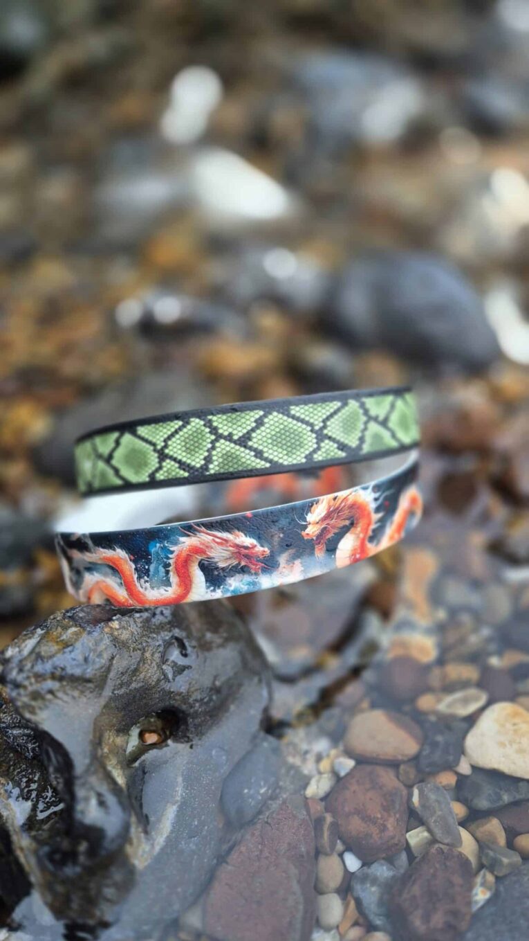 Three handmade BioThane dog collars stacked on a stone, featuring vibrant green geometric, white, and red dragon patterns.