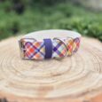 Handmade BioThane dog collar with tartan pattern in red, blue, orange and white, featuring silver hardware and navy webbing detail on wooden surface.