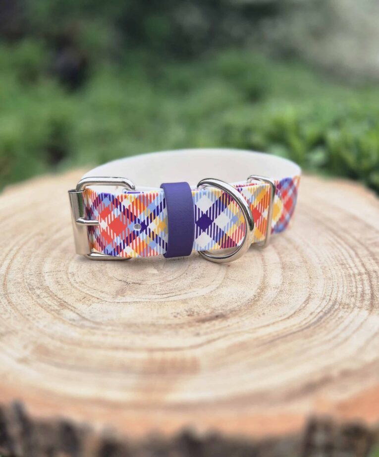 Handmade BioThane dog collar with tartan pattern in red, blue, orange and white, featuring silver hardware and navy webbing detail on wooden surface.