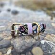 Handmade BioThane waterproof floral dog collar with purple trim and brass hardware, displayed on coastal pebbles.