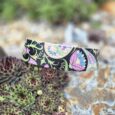 Vibrant handmade BioThane dog collar with intricate floral and botanical patterns in pink, blue, green and black against natural garden stones.
