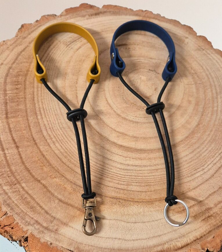 Two handmade BioThane wrist straps, lanyards with yellow and navy headbands, black cords, and metal hardware on natural wood.