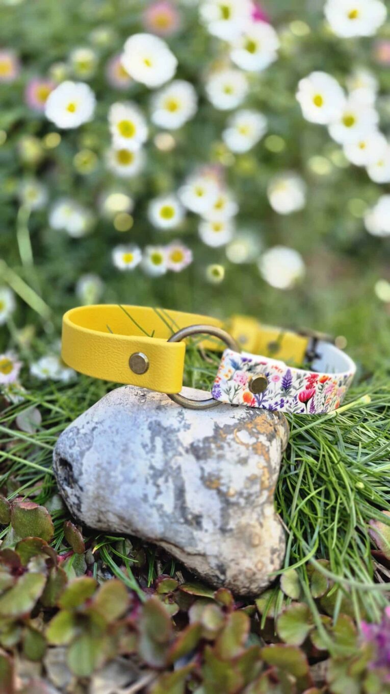 Yellow and floral printed handmade BioThane dog collars displayed on a stone in a garden with white flowers.