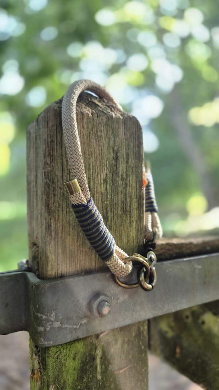 Handmade paracord dog collar with striped handle wrap, looped over weathered wooden post in garden setting.