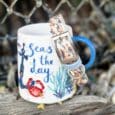 White mug with nautical design and blue handle, featuring a charm bracelet with sea-themed charms attached.