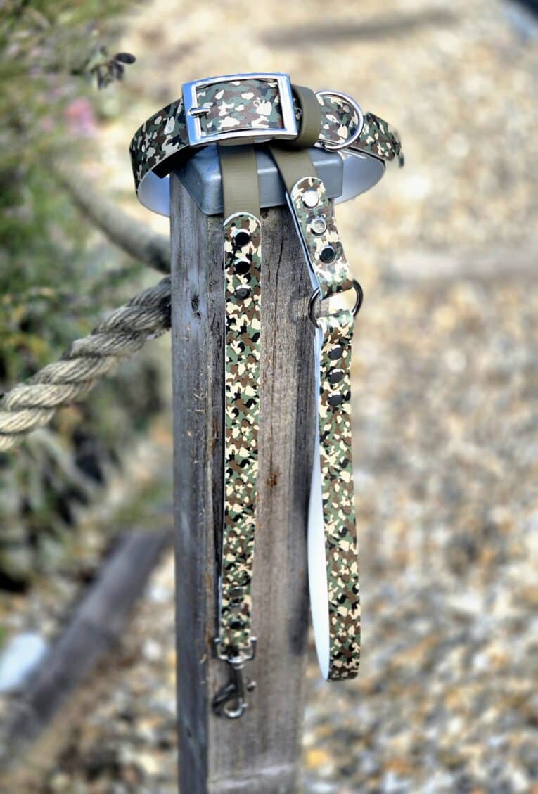 Handmade BioThane dog collars in camouflage pattern with blue buckle and metal D-ring displayed on wooden post.