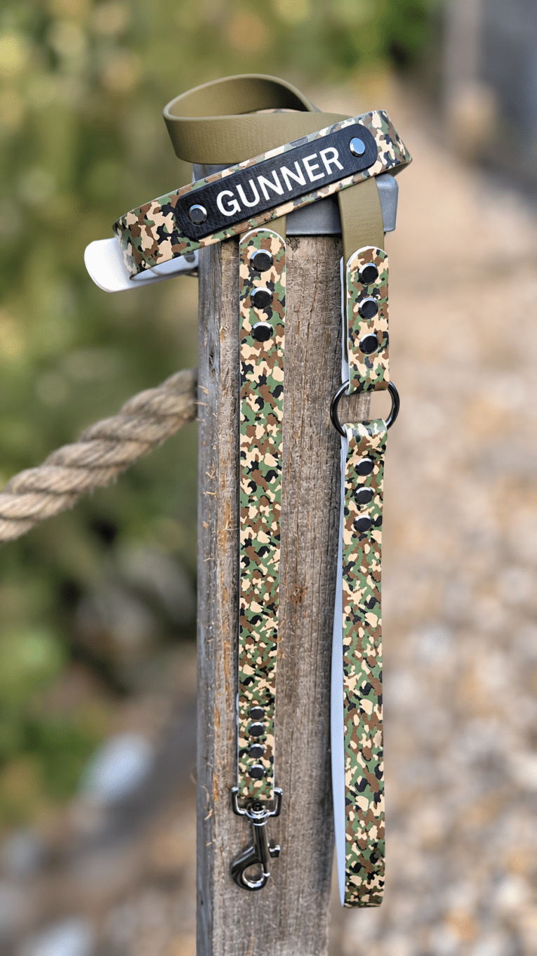 Camouflage handmade BioThane dog collar with 'GUNNER' label, displayed on wooden post with rope.