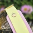 Handmade BioThane dog collar in soft yellow with pink binding and metal hardware, displayed outdoors among flowering plants.