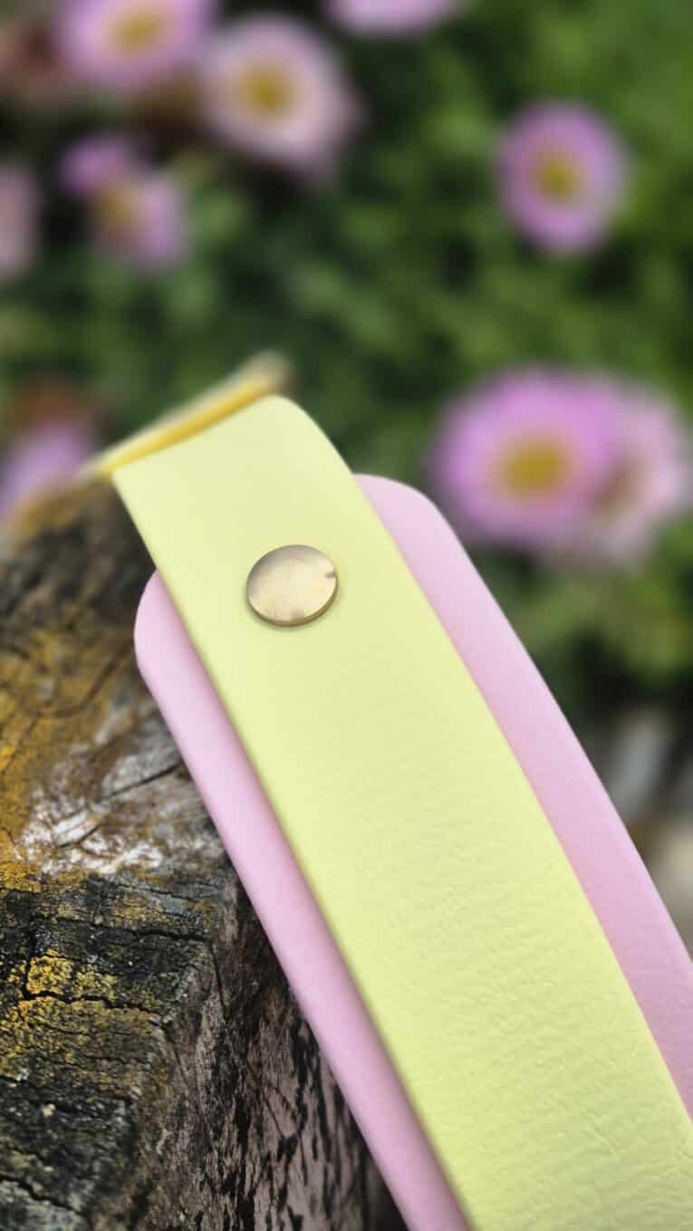Handmade BioThane dog collar in soft yellow with pink binding and metal hardware, displayed outdoors among flowering plants.