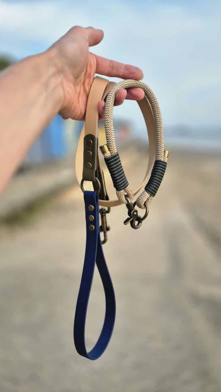 Hand holding a handmade paracord dog collar and biothane lead set in tan and navy, displayed on a beach.