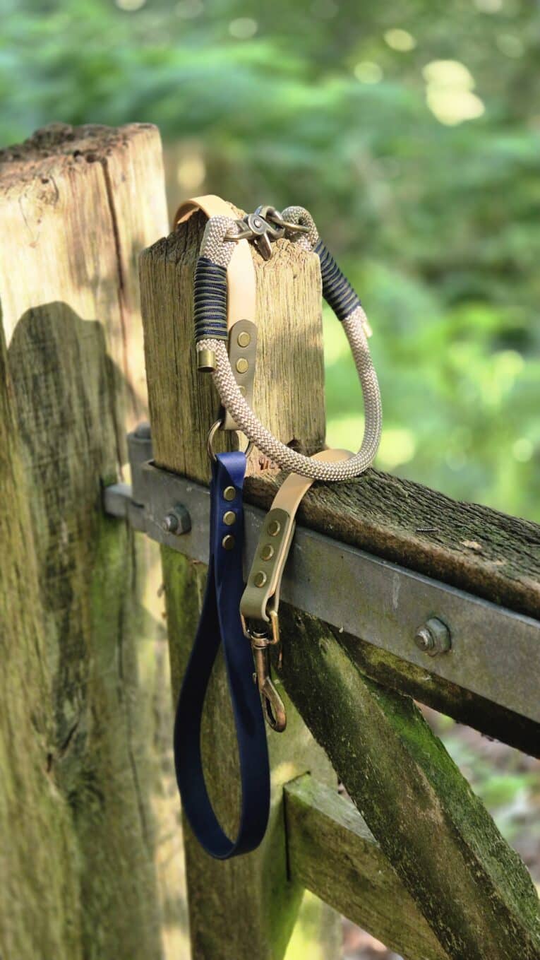 Handmade paracord dog collar and biothane lead in navy and tan, displayed on rustic wooden fence in countryside setting.